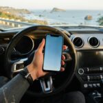 Hombre usando smartphone dentro de su coche junto al mar, gestionando seguro coche digital de manera online